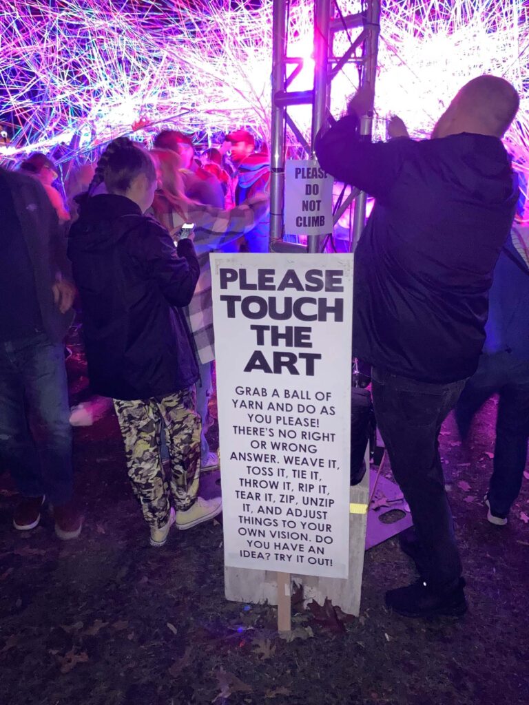 a crowd gathered around a string art installation with a sign that reads Please Touch the Art